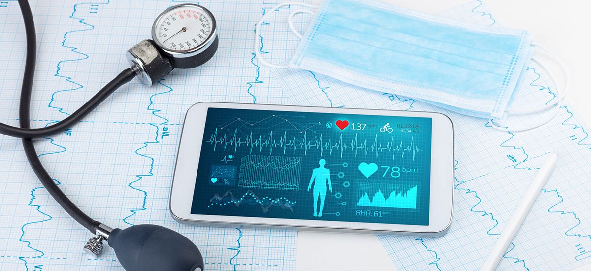 Image of a  blood pressure measuring device and a smartphone showing  vital signs on a background of a face mask and heart trace print-outs